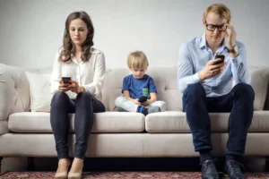 Pais e criança sentados no sofá cada um com o celular na mão